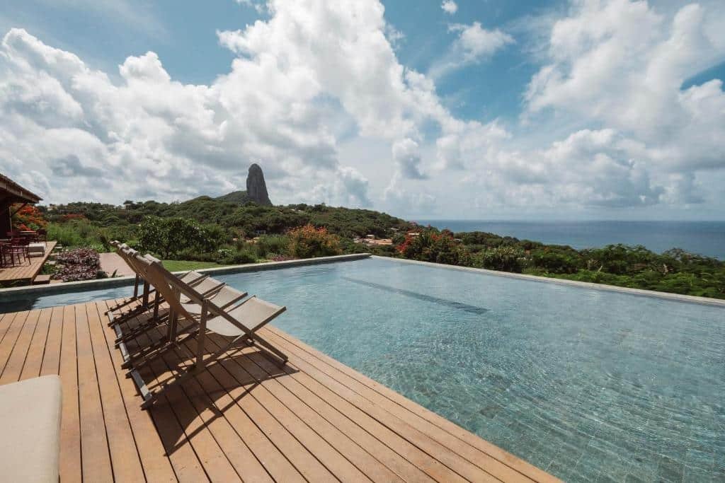 Piscina com deck e cadeiras na Colina Pousada Spa, uma das pousadas em Fernando de Noronha, com vista para o Morro do Pico e para o mar, em dia com muitas nuvens no céu