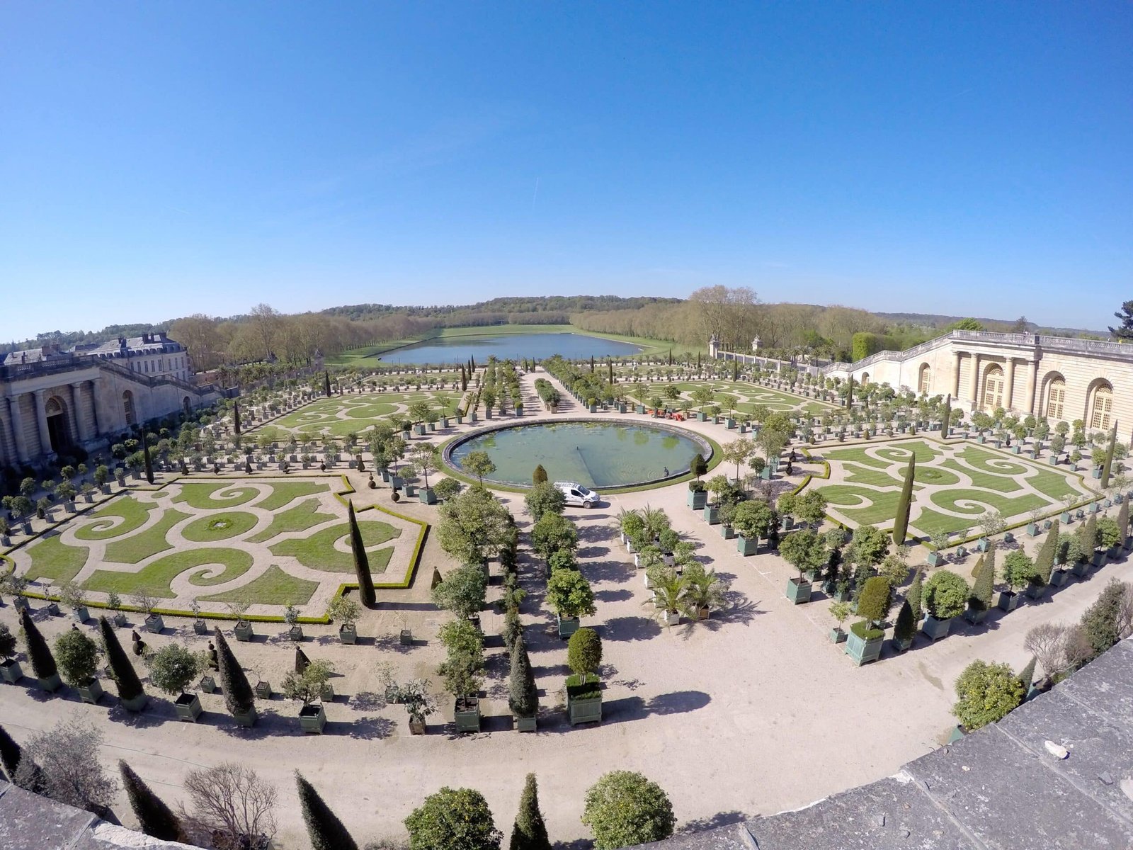 Jardins de Versalhes no Palácio de Versalhes com uma fonte circular no centro e várias árvores e jardins em volta aparados em formas geométricas. No fundo há um lago.
