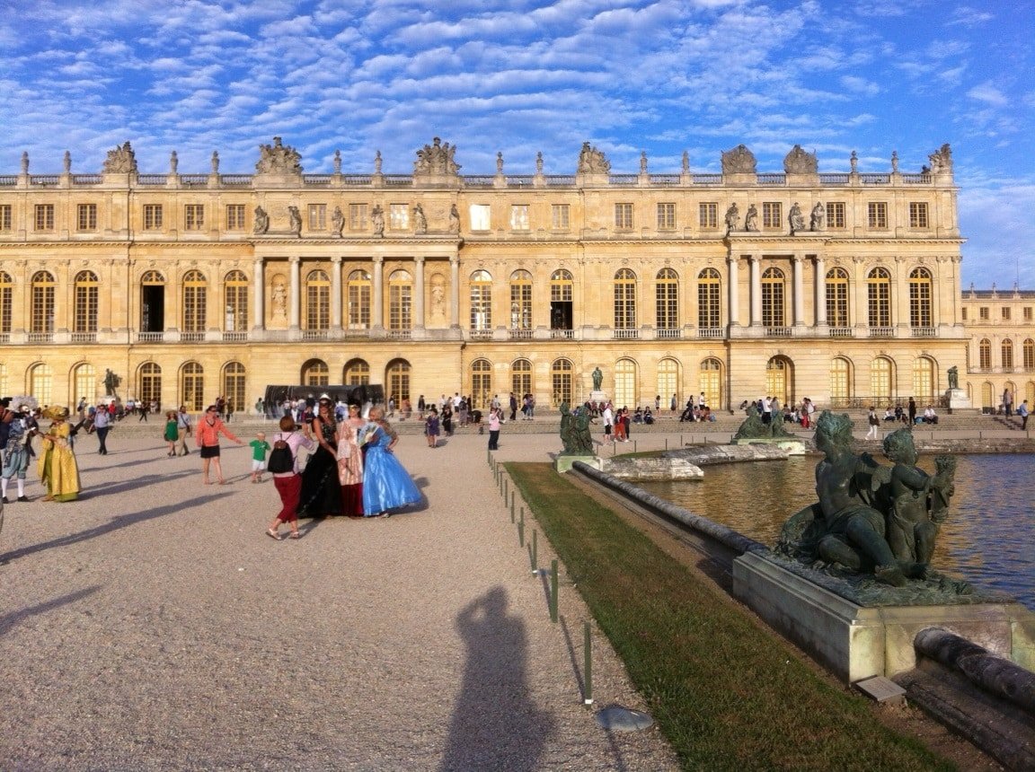 Palácio de Versalhes em um dia de céu azul com várias pessoas no pátio à frente. Na direita, há uma fonte larga com estátuas de pedra nas margens.