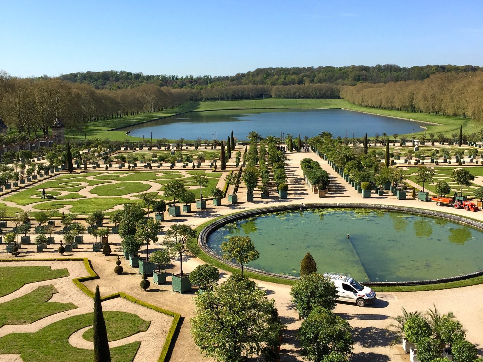 Jardins de Versalhes no Palácio de Versalhes com uma fonte circular no centro e várias árvores e jardins em volta aparados em formas geométricas. No fundo há um lago.