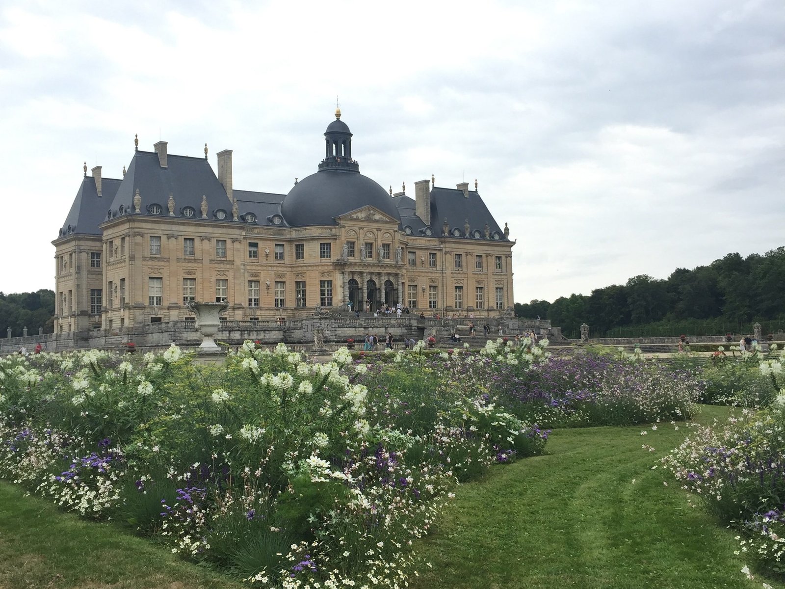 Foto do castelo Vaux-Le-Vimconte, antigra propriedade de Nicolas Fouquet. Há um vasto jardim florido na frente do castelo, que está ao fundo da foto.