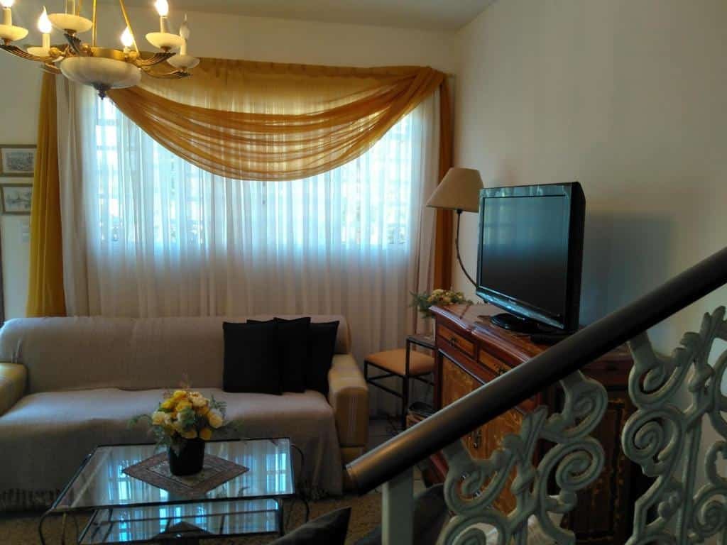 Sala em Condomínio Vila Paradiso. No canto inferior direito vemos de relance o corrimão da escada. Na frente está a sala com um sofá, uma mesa de centro de vidro e uma estante com TV encostada na parede à direita. Há uma janela com cortina atrás do sofá.
