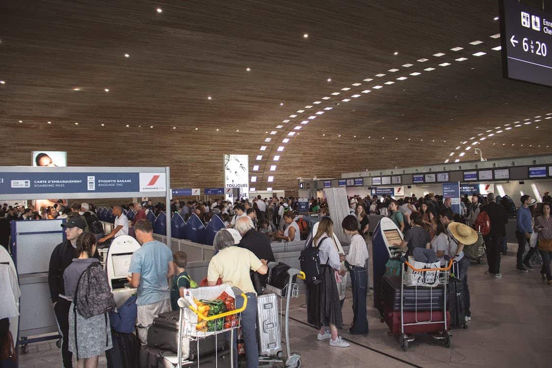 Um terminal de aeroporto lotado com pessoas esperando na fila nos balcões de check-in. Carrinhos de bagagem e placas são visíveis. A imagem ilustra o post sobre como chegar em Paris.