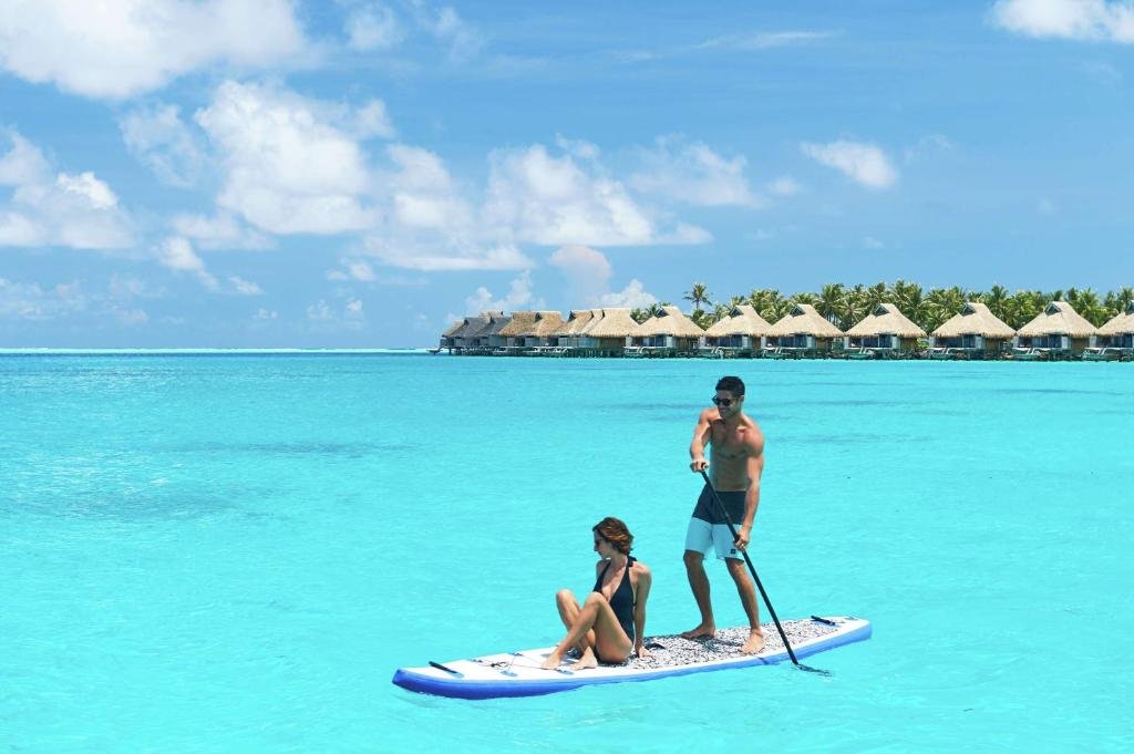 Um homem está de pé praticando paddleboard enquanto uma mulher está sentada na prancha. Eles estão em águas azuis claras com bangalôs sobre a água à distância.