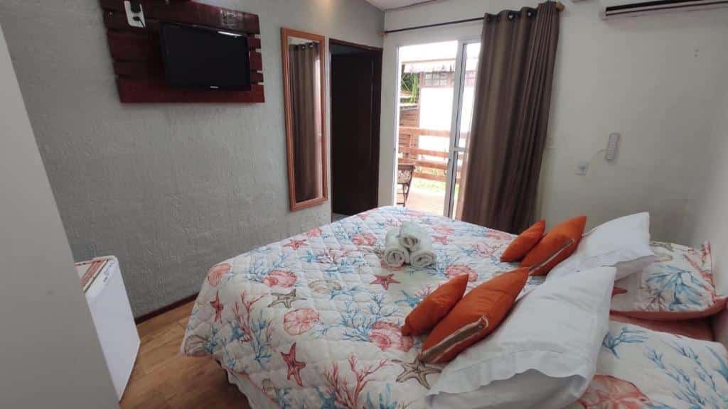 Quarto de casal na Corrente Marítima Pousada, com uma cama com edredom de motivos marítimos, uma cortinha em frente à porta de entrada, ar-condicionado, espelho de corpo inteiro, frigobar e TV
