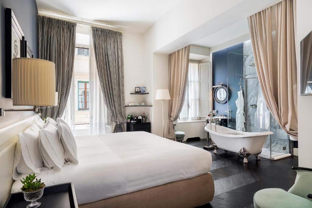 Quarto do Corte Calzaiuoli Elegant Suites, com cama de casal com travesseiros em cima, janela com cortina grande cinza, e banheiro integrado com jacuzzi e um box de vidro do chuveiro