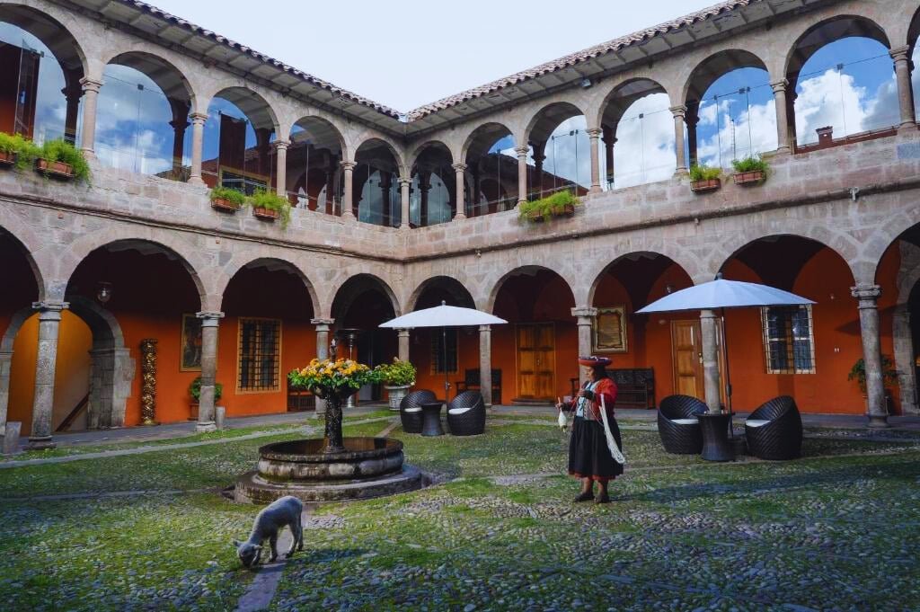 pátio aberto do hotel Costa del Sol Wyndham em Cusco com uma fonte de pedra ao centro e arcos de pedra nas paredes atrás. Há também um cordeinrinho branco e uma mulher com roupas típicas peruanas.