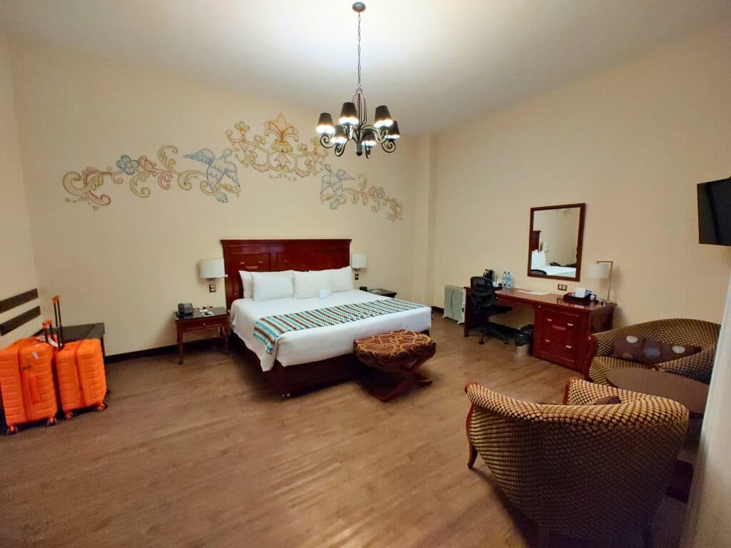 quarto do hotel Costa del Sol Wyndham Cusco com uma cama de casal ao centro, um espaço para trabalho à direita, duas poltronas em frente a cama, e uma área com bancos e malas à direita.