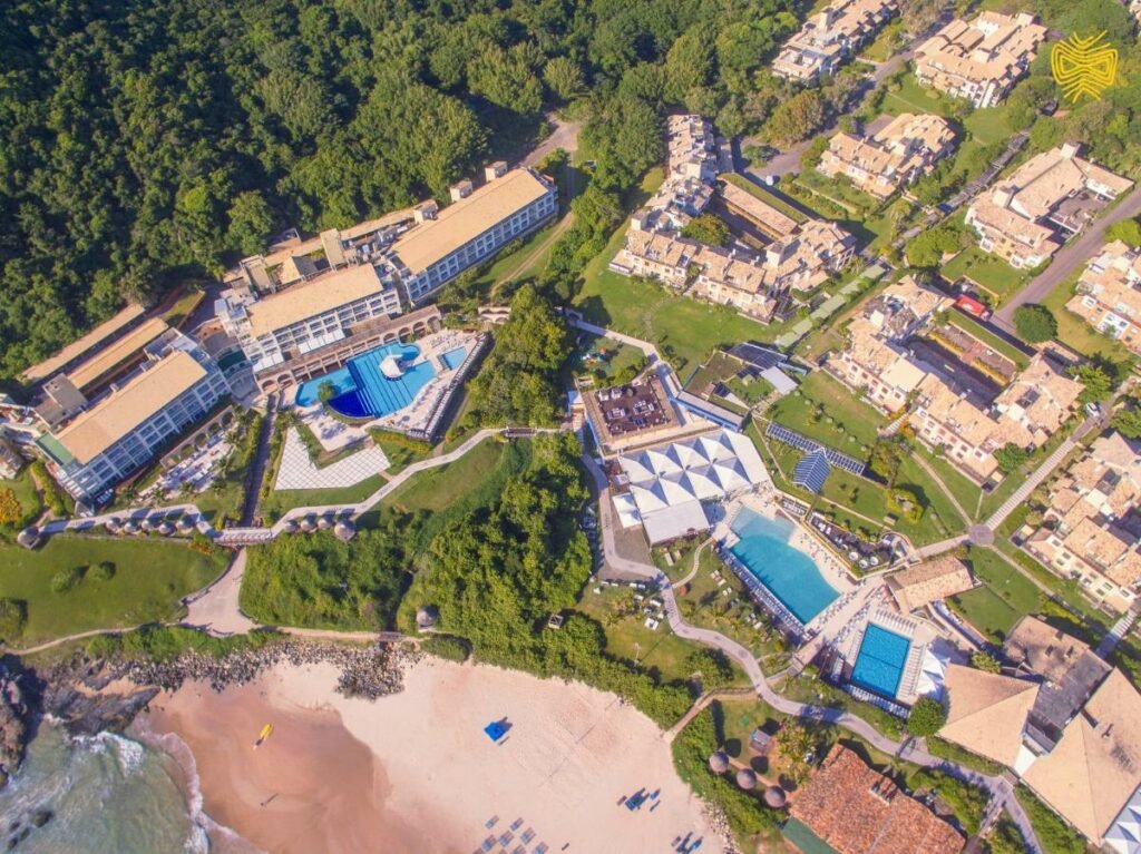 A imagem aérea do Costão do Santinho Resort All Inclusive mostra uma extensa propriedade à beira-mar, rodeada por uma densa vegetação. O resort inclui várias piscinas, áreas de lazer, e edificações para hospedagem distribuídas harmoniosamente pelo terreno. Ao fundo, há uma praia com areia clara, onde algumas cadeiras e guarda-sóis estão dispostos. As instalações do resort parecem bem integradas à paisagem natural, oferecendo uma vista impressionante do oceano e das áreas verdes circundantes.