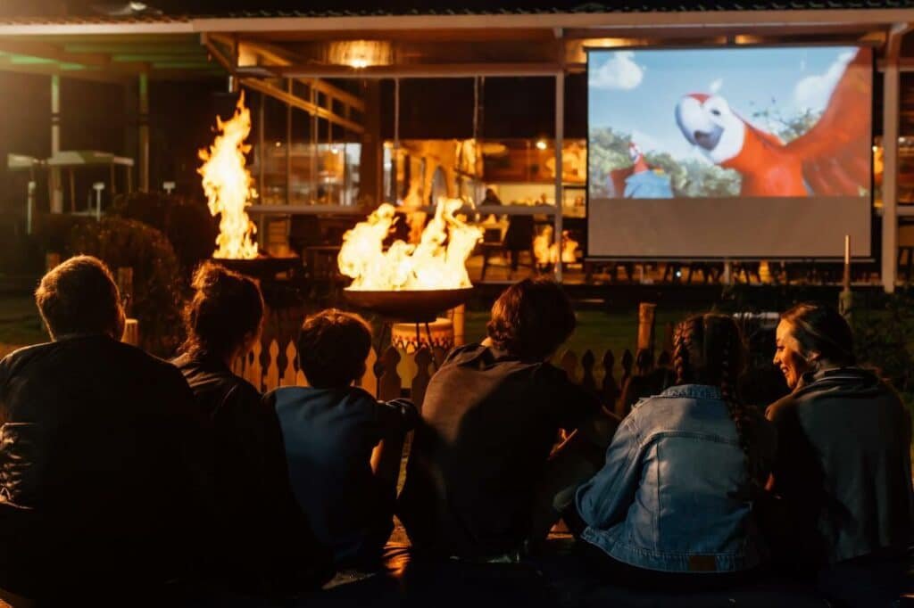 A imagem mostra uma sessão de cinema ao ar livre no Costão do Santinho Resort All Inclusive, um dos melhores resorts all inclusive no Brasil. Várias pessoas estão sentadas em bancos assistindo a um filme projetado em uma tela grande. Em primeiro plano, há duas tochas acesas, criando uma atmosfera acolhedora e iluminada. Ao fundo, o filme exibe uma cena com um papagaio colorido.