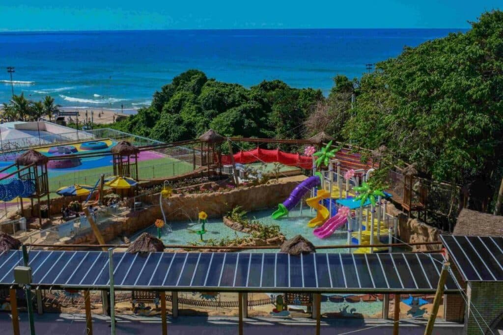 A imagem mostra uma área de recreação infantil no Costão do Santinho Resort All Inclusive. Há várias estruturas coloridas de playground, incluindo tobogãs e brinquedos aquáticos, em uma piscina rasa. Ao fundo, pode-se ver uma vista panorâmica do oceano com ondas suaves e uma faixa de areia clara. A área está rodeada por vegetação exuberante, criando um ambiente tropical e vibrante. O céu está claro e azul, sugerindo um dia ensolarado.