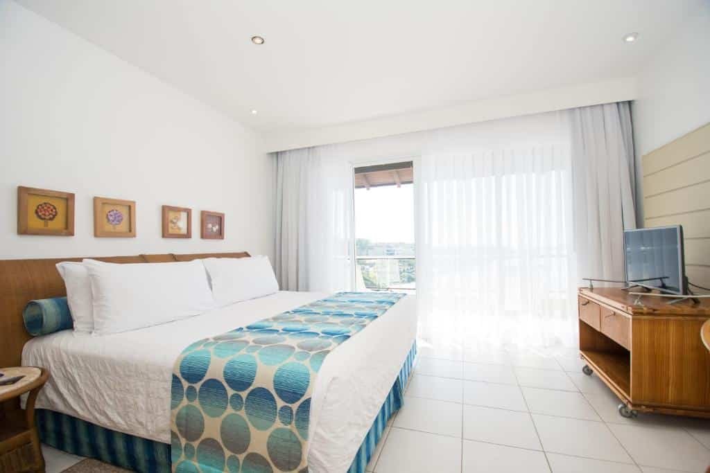 A imagem mostra um quarto do Costão do Santinho Resort All Inclusive, um dos melhores resorts all inclusive no Brasil. A cama de casal, com lençóis brancos e uma manta decorativa em tons de azul, ocupa o centro da cena. Há quatro quadros com flores na parede acima da cama. À direita, uma grande janela com cortinas brancas translúcidas deixa entrar bastante luz natural, revelando uma varanda com vista para a área externa. Um móvel de madeira com uma TV está posicionado à frente da cama. O ambiente é claro e decorado de forma simples e elegante, transmitindo uma sensação de conforto e tranquilidade.