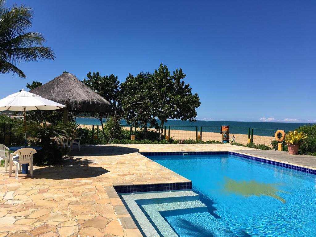 Piscina do O Costão do Sol, uma das pousadas em Balneário Camboriú, com algumas mesas com guarda-sóis ao lado e a praia logo ao lado, com areia bege e mar azul escuro em um dia ensolarado e com poucas nuvens