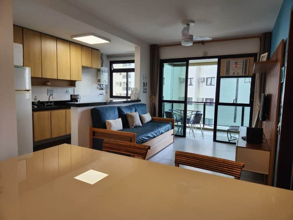 Sala de estar do Apartamento Barra Pé na Areia com mesa a frente com cadeiras do lado direito da imagem sofá com uma cômoda com TV e atrás do sofá um balcão e ao fundo armário da cozinha e geladeira.