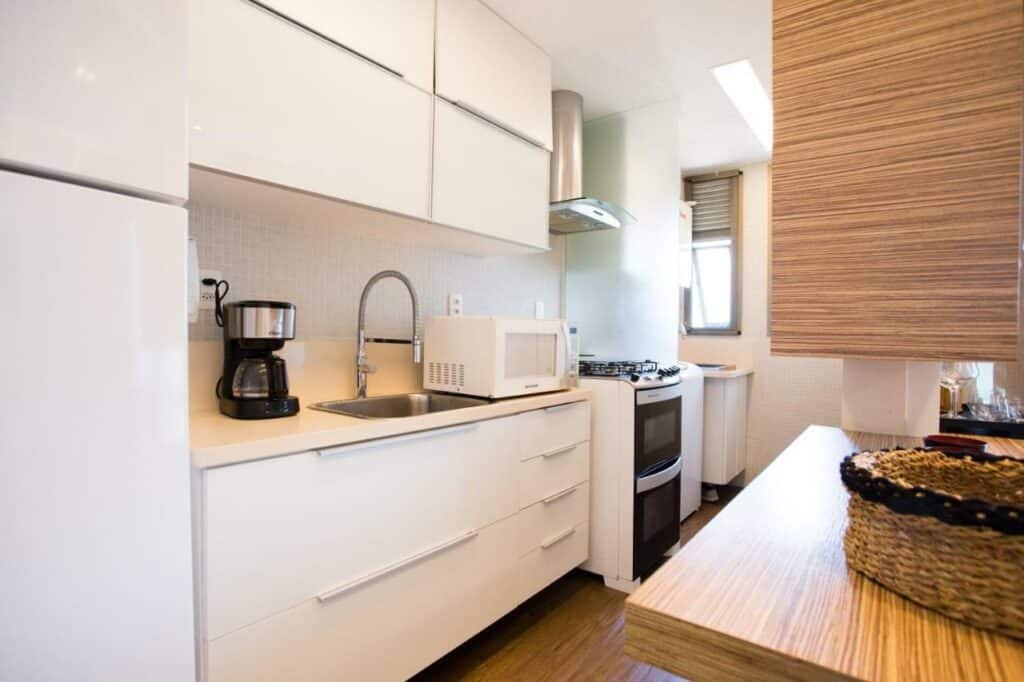 Cozinha do Barra da Tijuca de frente para o mar – 2 suítes até 6 pessoas em camas com mesa do lado direito da imagem e do lado esquerdo armário com pia, micro-ondas e fogão.