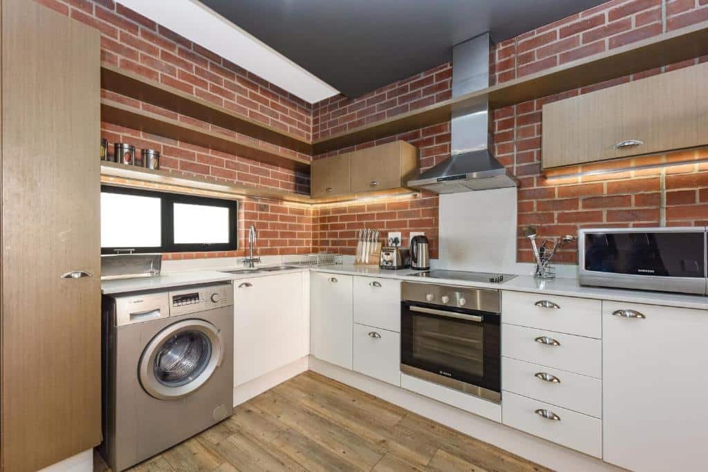 Cozinha moderna do Easy Stay - The Vantage Rosebank com paredes de tijolos, armários brancos, eletrodomésticos de aço inoxidável e uma máquina de lavar roupa sob o balcão.