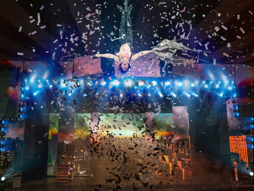 Criss Angel suspenso de cabeça para baixo sobre um palco em meio a confetes, com luzes brilhantes e uma paisagem urbana ao fundo.