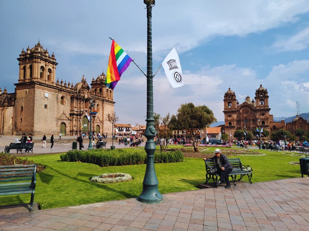 Plaza de las armas de Cusco com um poste de luz bem ao centro, com duas bandeiras de cada lado: a inca e a peruana. Ao fundo é possível ver a Catedral de Cusco à esquerda e a Iglesia de la compañia de Jesus à direita.