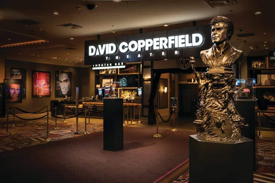 Entrada do Teatro David Copperfield, com uma grande estátua e letreiros iluminados, com um bar de teatro ao fundo.