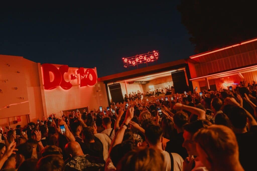 Uma grande multidão se reúne ao ar livre à noite, de frente para um palco com um DJ. Letreiros de neon vermelho e luzes iluminam a cena na DC-10, uma das festas em Ibiza.