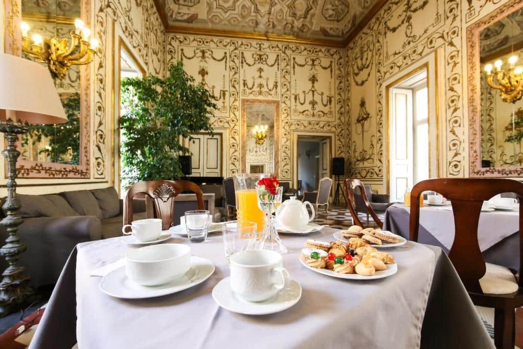 Mesa do café da manhã do Decumani Hotel De Charme com algumas guloseimas em cima. Em volta há outras mesas parecidas, um sofá no canto esquerdo perto de uma planta verde e a decoração do ambiente é antiga e clássica. Imagem ilustrando post Hotéis em Nápoles.