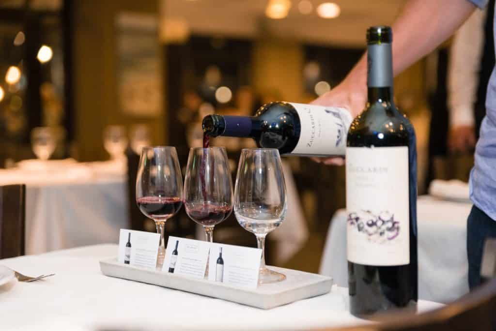 Na imagem vemos três taças postas em uma pequena bandeja, com plaquinhas indicativas de vinho à frente. A taça do meio está sendo serviça com vinho ZuccardiQ. Ao lado há outra garrafa de vinho Zuccardi. Foto da página da bodega no Facebook. Vinicola Mendoza