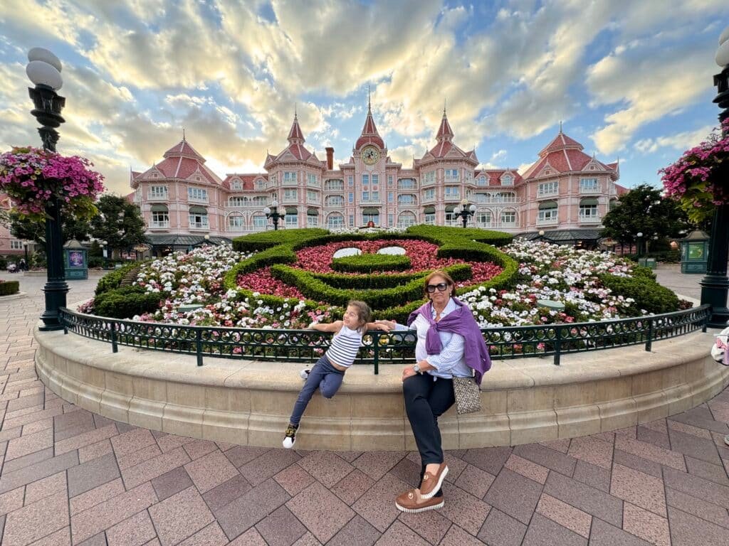 Uma mulher e uma criança estão sentadas em um muro baixo em frente a um grande arranjo floral do Mickey Mouse e a um edifício rosa em estilo vitoriano. Esse é um dos pontos turísticos de Paris.