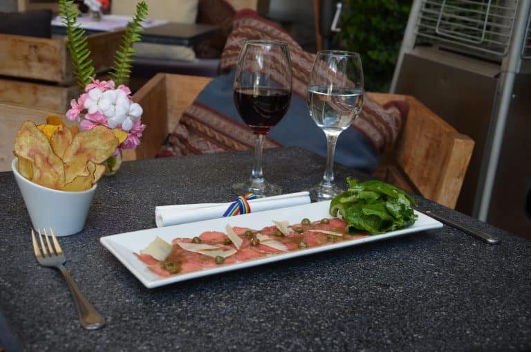 Uma mesa do Divina Comédia Restaurante com duas taças de vinho, um prato retangular de carne, salada e uma flor ao lado de uma xícara de batatas fritas.