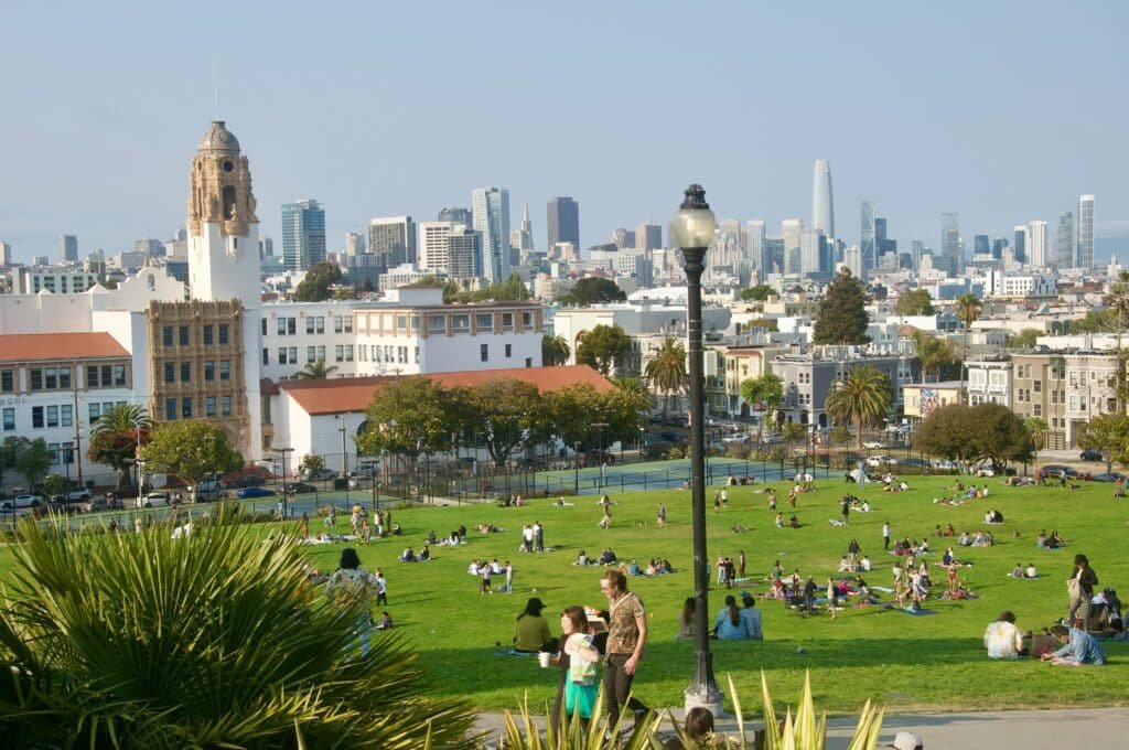 Foto de um pedaço do gramado em Dolores Park, onde diversas pessoas estão sentadas e deitadas. Ao fundo, do lado de fora do parque, estão várias construções.
