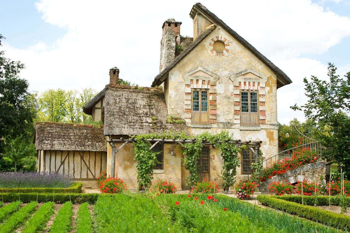Uma casa rústica com hera, telhado de palha e um jardim de flores ao redor em um dia ensolarado. É o domínio de Maria Antonieta no Palácio de Versalhes.
