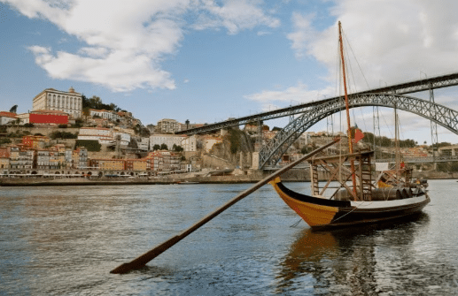 Um barco de madeira tradicional flutua em um rio com uma cidade e uma ponte metálica em arco ao fundo, sob um céu nublado.