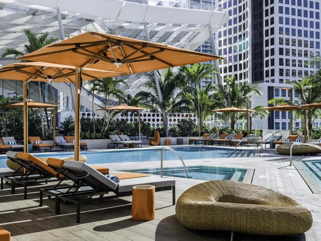 A imagem mostra a área da piscina no East Miami, um dos hotéis em Miami Beach indicados. Há espreguiçadeiras com almofadas laranjas e brancas ao redor da piscina, cobertas por grandes guarda-sóis laranja. Atrás das espreguiçadeiras, há vegetação tropical com palmeiras e arbustos. Em segundo plano, grandes edifícios modernos de vidro e metal compõem o cenário, sugerindo uma localização central em uma cidade grande. A atmosfera é sofisticada e relaxante, ideal para lazer.