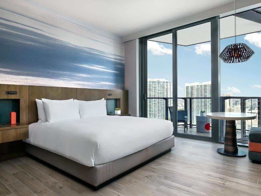 A imagem mostra um quarto do East Miami com uma cama de casal grande, coberta com lençóis brancos. A parede atrás da cama tem um mural com uma imagem de praia, criando uma atmosfera relaxante. O quarto possui piso de madeira clara e uma grande janela de vidro que leva a uma varanda, oferecendo uma vista de prédios altos e o céu azul. Há uma mesa redonda com cadeiras na varanda e uma luminária pendente moderna ao lado da cama.