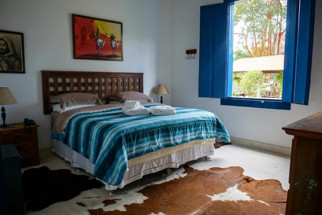 Quarto com uma cama de casal coberta com um cobertor listrado azul, tapete de couro no chão, arte na parede e uma janela aberta com venezianas azuis no Hotel Fazenda Bela Riba, um dos hotéis fazenda no Rio de Janeiro.