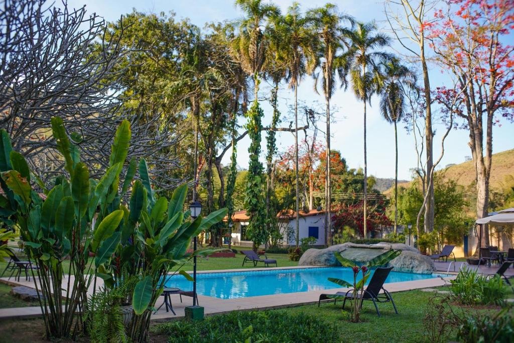 Piscina ao ar livre cercada por espreguiçadeiras, plantas tropicais e palmeiras altas sob um céu azul claro no Hotel Fazenda Bela Riba.