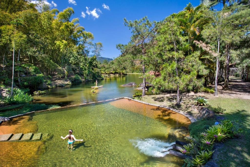 Eco Resort Hotel Villa São Romão em Lumiar, com piscina natural em frente à lago e muita área verde, em um dia de céu azul intenso com poucas nuvens.