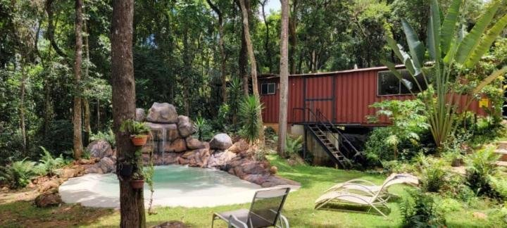 Container vermelho com uma escada dando acesso ao interior do Econtainer Suítes Macacos, com bastante natureza verde em volta. No lado esquerdo há uma piscina com pedras e cascata caindo água, além de algumas espreguiçadeiras em volta para ilustrar as pousadas em Macacos.