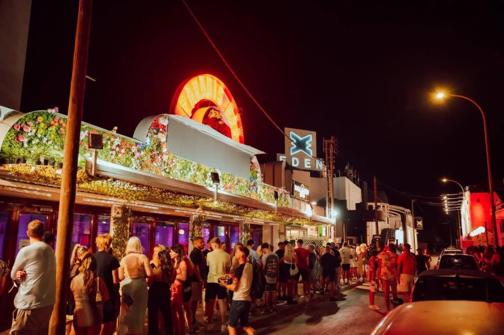 Pessoas esperando na fila do lado de fora de uma boate à noite, com letreiros de neon e entrada decorada visíveis. Essa é a Eden Ibiza, uma das melhores festas em Ibiza.