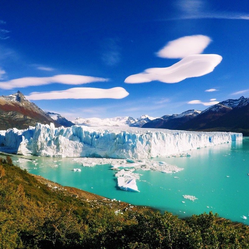 Geleira Perito Moreno com nuvens lenticulares em um céu azul claro e montanhas ao fundo.