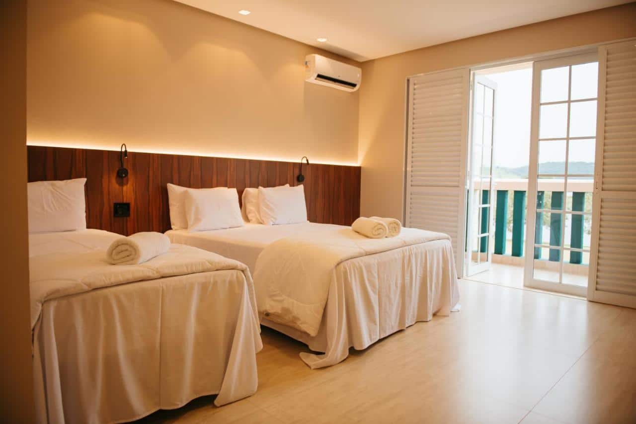 Um quarto do Engenho da Serra Hotel EcoResort com duas camas bem arrumadas, lençóis brancos, luminárias de parede, ar condicionado e uma varanda com portas abertas.