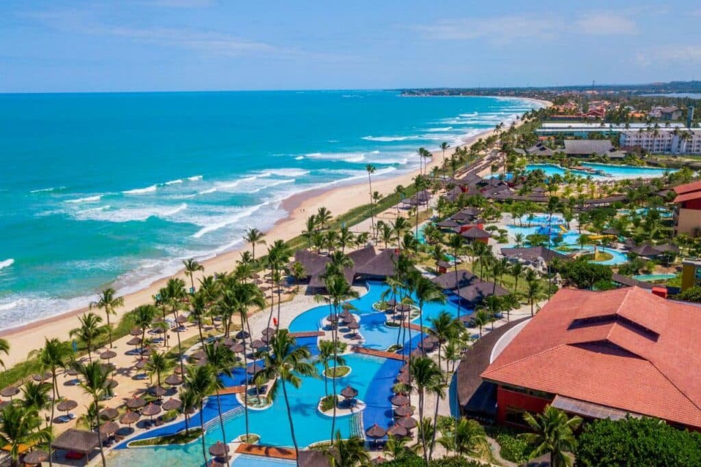 A imagem aérea mostra o Enotel Porto de Galinhas All Inclusive, localizado à beira-mar, com uma vista panorâmica deslumbrante do oceano azul-turquesa e a extensa praia de areia branca. O resort é cercado por uma exuberante vegetação tropical e possui várias piscinas sinuosas, intercaladas com áreas de descanso com guarda-sóis e espreguiçadeiras. À direita, destaca-se um grande prédio com telhado vermelho, provavelmente a área principal do resort.