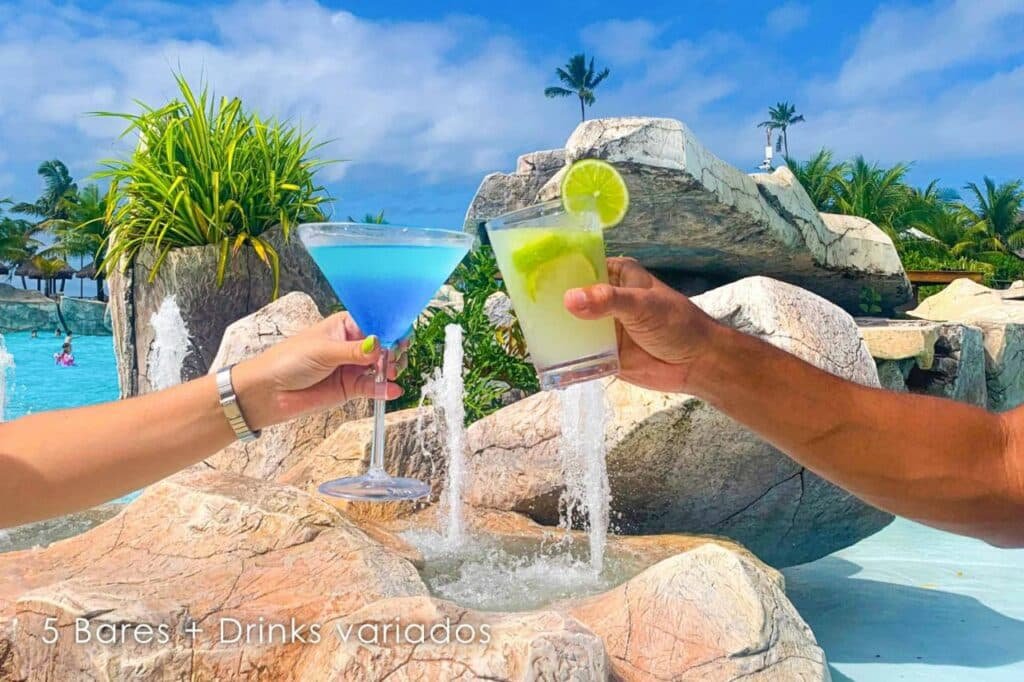 A imagem mostra duas pessoas brindando com drinks coloridos em um ambiente ao ar livre do Enotel Porto de Galinhas All Inclusive, um dos melhores resorts all inclusive no Brasil. À esquerda, um drink azul em uma taça de coquetel é segurado por uma mão feminina, e à direita, um drink verde com uma fatia de limão é segurado por uma mão masculina. Ao fundo, há uma piscina com cascatas de pedras artificiais e vegetação tropical, criando uma atmosfera relaxante e paradisíaca. A legenda na imagem menciona "5 Bares + Drinks variados", indicando a variedade de opções de bebidas oferecidas no local.