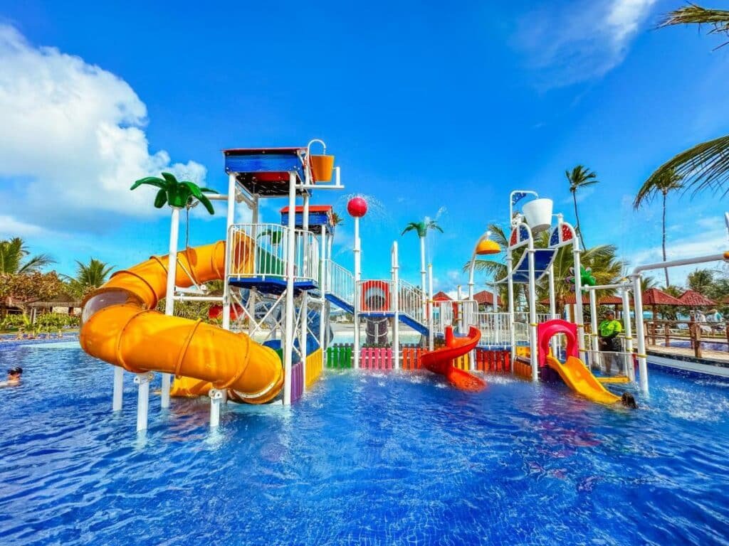 A imagem mostra um playground aquático em uma piscina, com várias estruturas coloridas e escorregadores para crianças no Enotel Porto de Galinhas All Inclusive. À esquerda, há um escorregador tubular laranja e, ao centro, há dois escorregadores abertos, um vermelho e um amarelo. No topo das estruturas, há decorações que lembram palmeiras e baldes que derramam água periodicamente, criando um ambiente divertido e interativo. O céu azul com algumas nuvens e a vegetação tropical ao fundo completam o cenário.