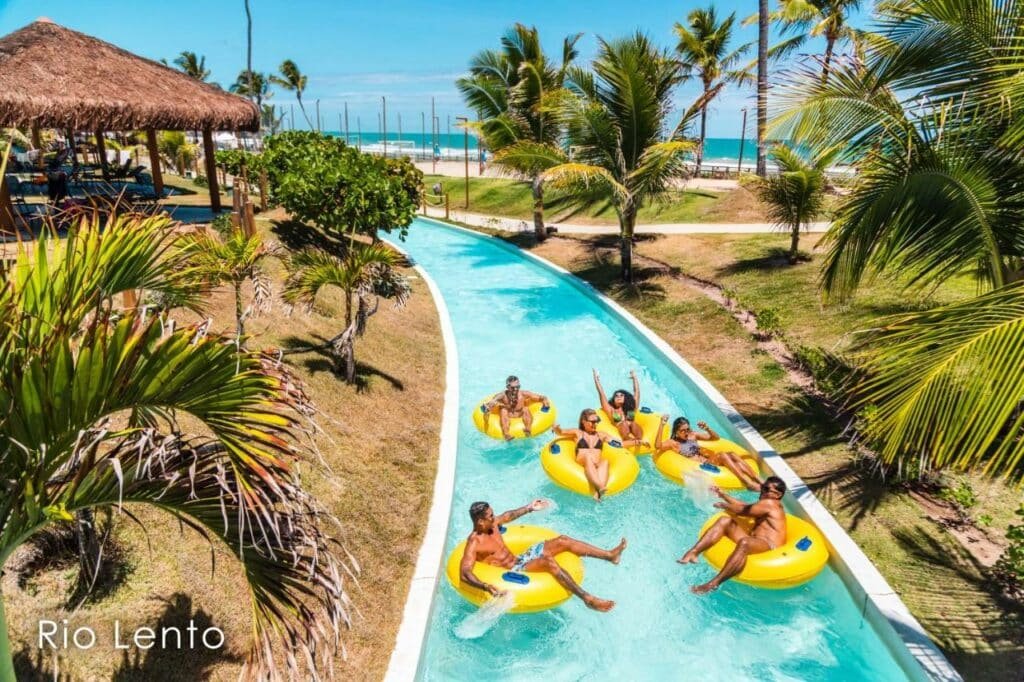 A imagem mostra um grupo de pessoas relaxando em bóias amarelas no rio lento do Enotel Porto de Galinhas All Inclusive, rodeado por palmeiras e vegetação tropical. O rio sinuoso passa por uma área verde bem cuidada e está próximo à praia, visível ao fundo com o mar azul. Há uma cabana de palha à esquerda, proporcionando sombra e um local de descanso. As pessoas estão sorrindo e aproveitando o ambiente ensolarado, evidenciando um clima descontraído e agradável no resort.