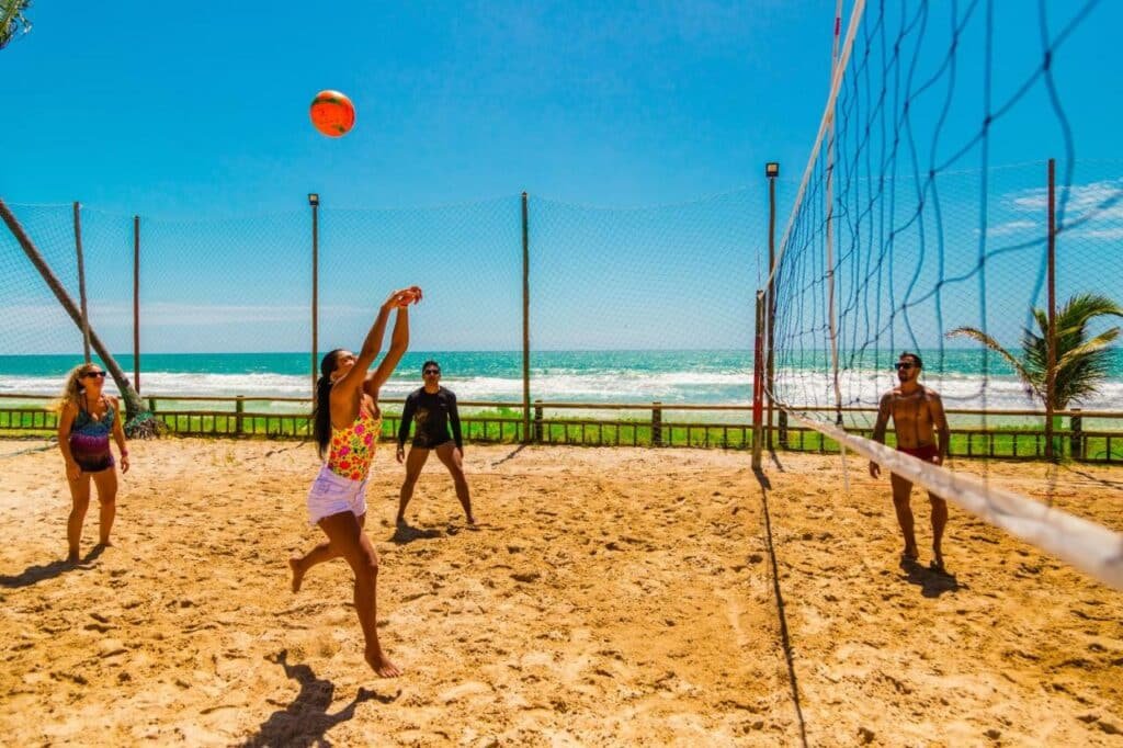 A imagem mostra quatro pessoas jogando vôlei de praia em uma quadra de areia cercada por uma rede de proteção, com o mar azul ao fundo, no Enotel Porto de Galinhas All Inclusive, um dos melhores resorts all inclusive no Brasil. Uma mulher está prestes a dar um toque na bola, enquanto os outros jogadores observam e se preparam para a jogada. O céu está claro e ensolarado, criando um ambiente vibrante e alegre.