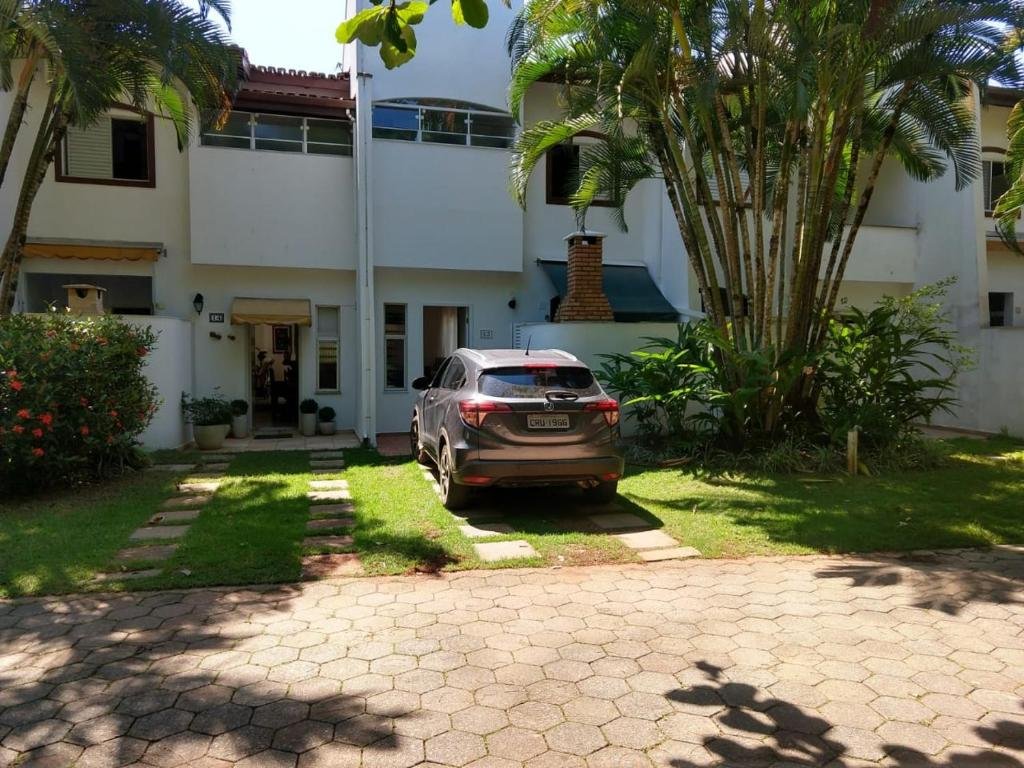 Entrada da Casa na Toninhas, uma construção pintada de branco, com dois andares e algumas árvores na frente, junto de um jardim, para representar pousadas na Praia das Toninhas