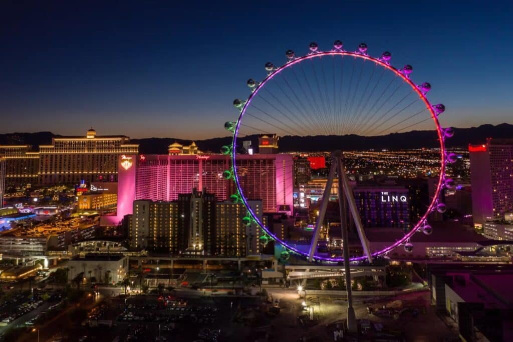 A imagem mostra a High Roller, uma roda-gigante toda iluminada durante à noite de Vegas. Ao fundo estão os hotéis e demais construções, também iluminados.