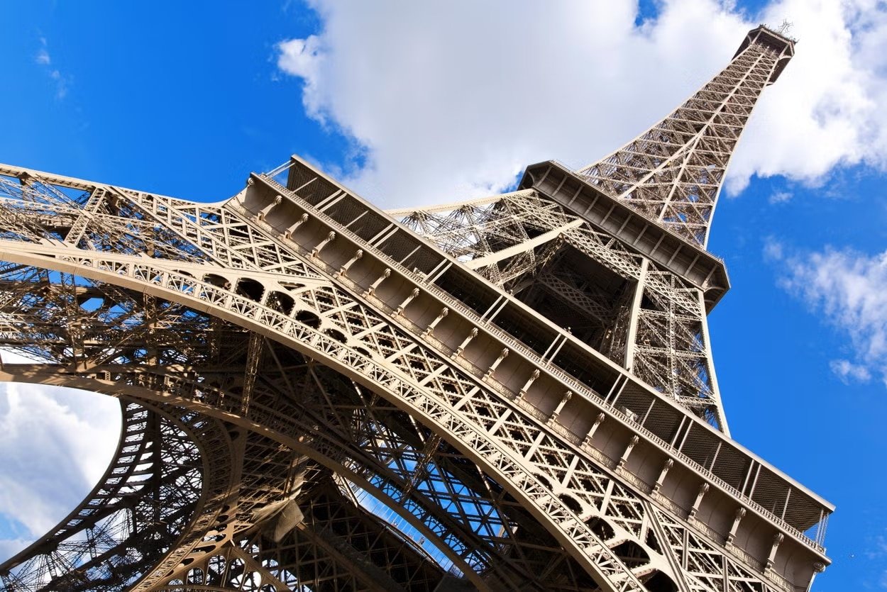 Vista de ângulo baixo da Torre Eiffel contra um fundo de céu azul e nuvens brancas para ilustrar o post sobre pontos turísticos de Paris.