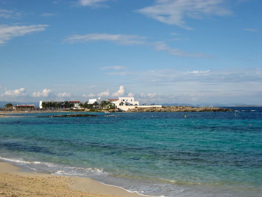 Vista de uma praia com areia fina, água azul-turquesa e um edifício branco ao longe sob um céu parcialmente nublado no vilarejo de Es Pujols, uma das opções de o que fazer em Formentera.