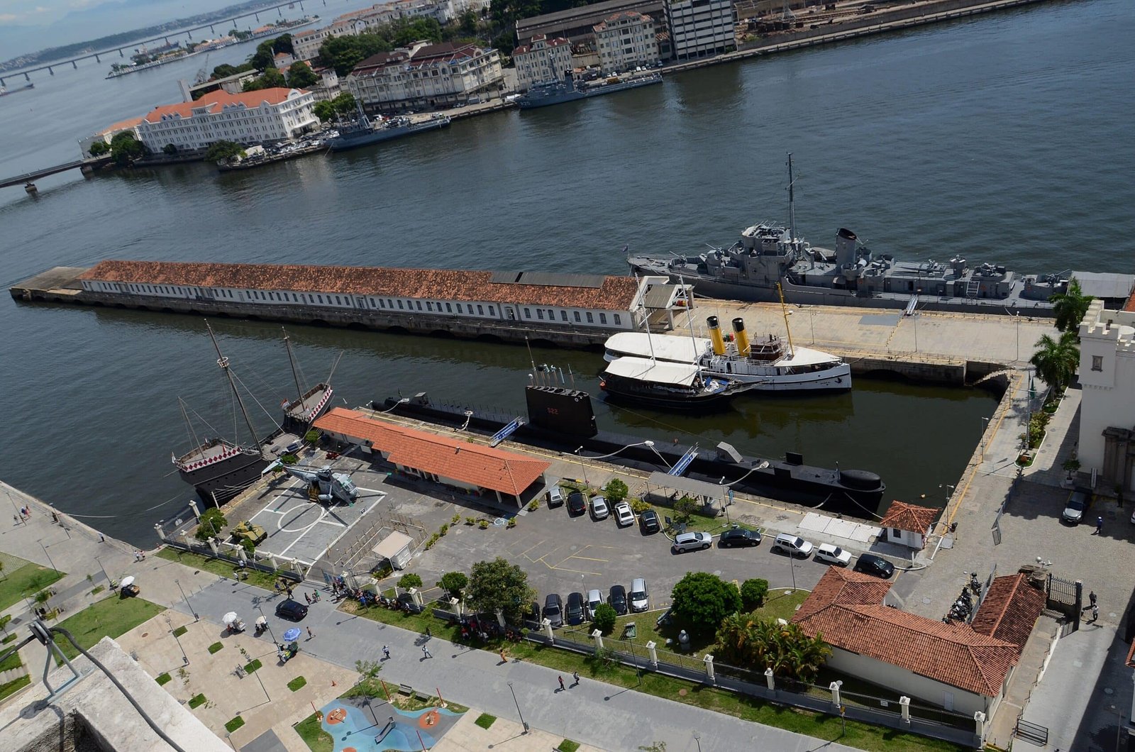 Vista aérea do Espaço Cultural da Marinha com um navio de guerra e edifícios, cercado por água e paisagem urbana ao fundo.